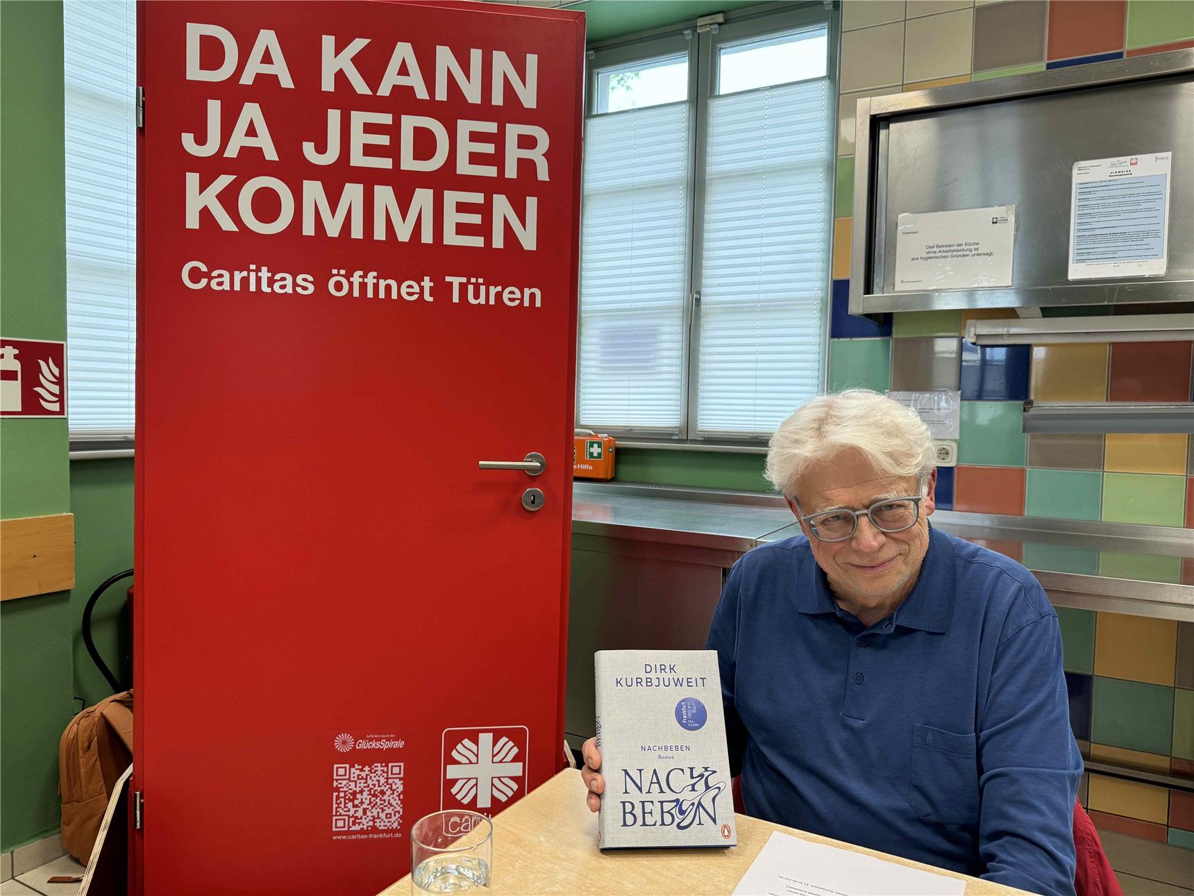 Der Autor, Sprecher und Regisseur Helge Heynold mit Dirk Kurbjuweits Roman 'Nachbeben'. Er sitzt vorne am Lesungspult neben der großen roten Caritas Tür.