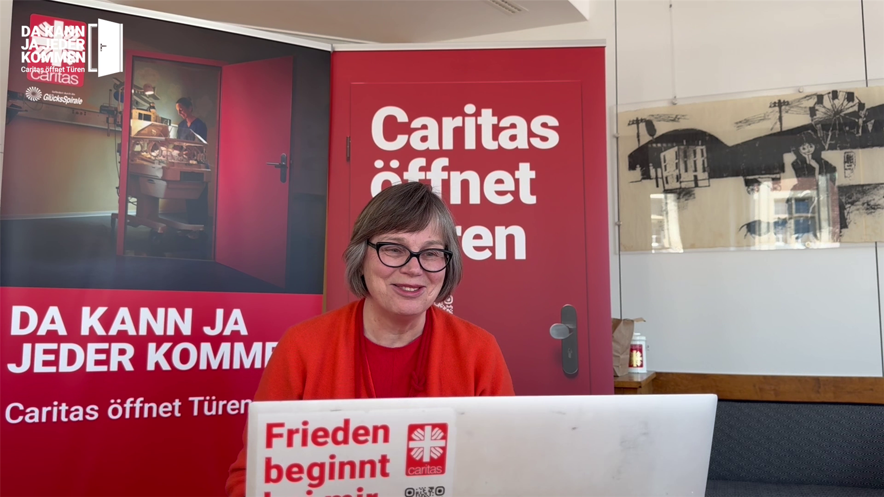 Die Präsidentin der Caritas sitzt lächelnd vor dem Computer und nimmt an einer Video-Konferenz teil. Im Hintergrund ist das Plakat der Kampagne zu sehen.