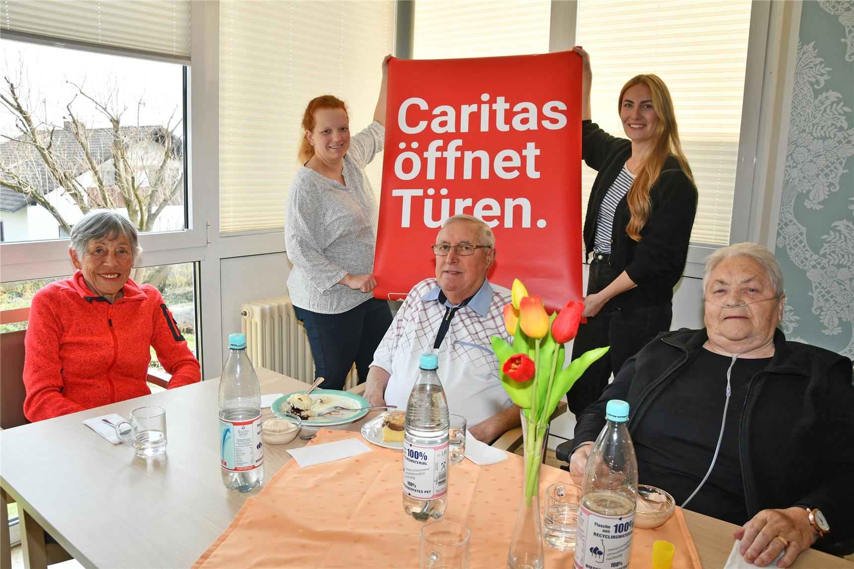 Die Leiterin des Seniorenheims Sarah Jacoby und Küchenleiterin Ute Ostermeier halten beim offenen Mittagstisch das Plakat mit der AUfschrift "Caritas öffnet Türen" hoch. Im Vordergrund sind drei Gäste des offnen Mittagstischs zu sehen.