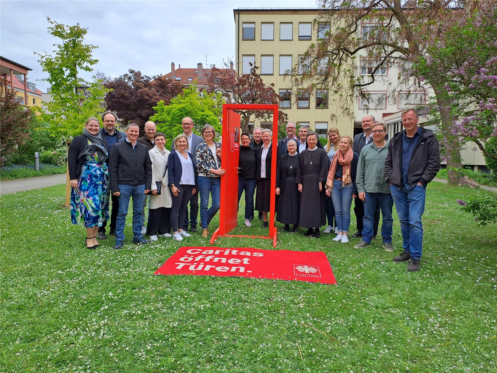 Eine Gruppe von ca. 20 Mitarbeiter*innen steht mit Fra Welskop-Deffaa au feiner blühenden Frühlingswise. Im Zentrum die große rote Kampagnentür.