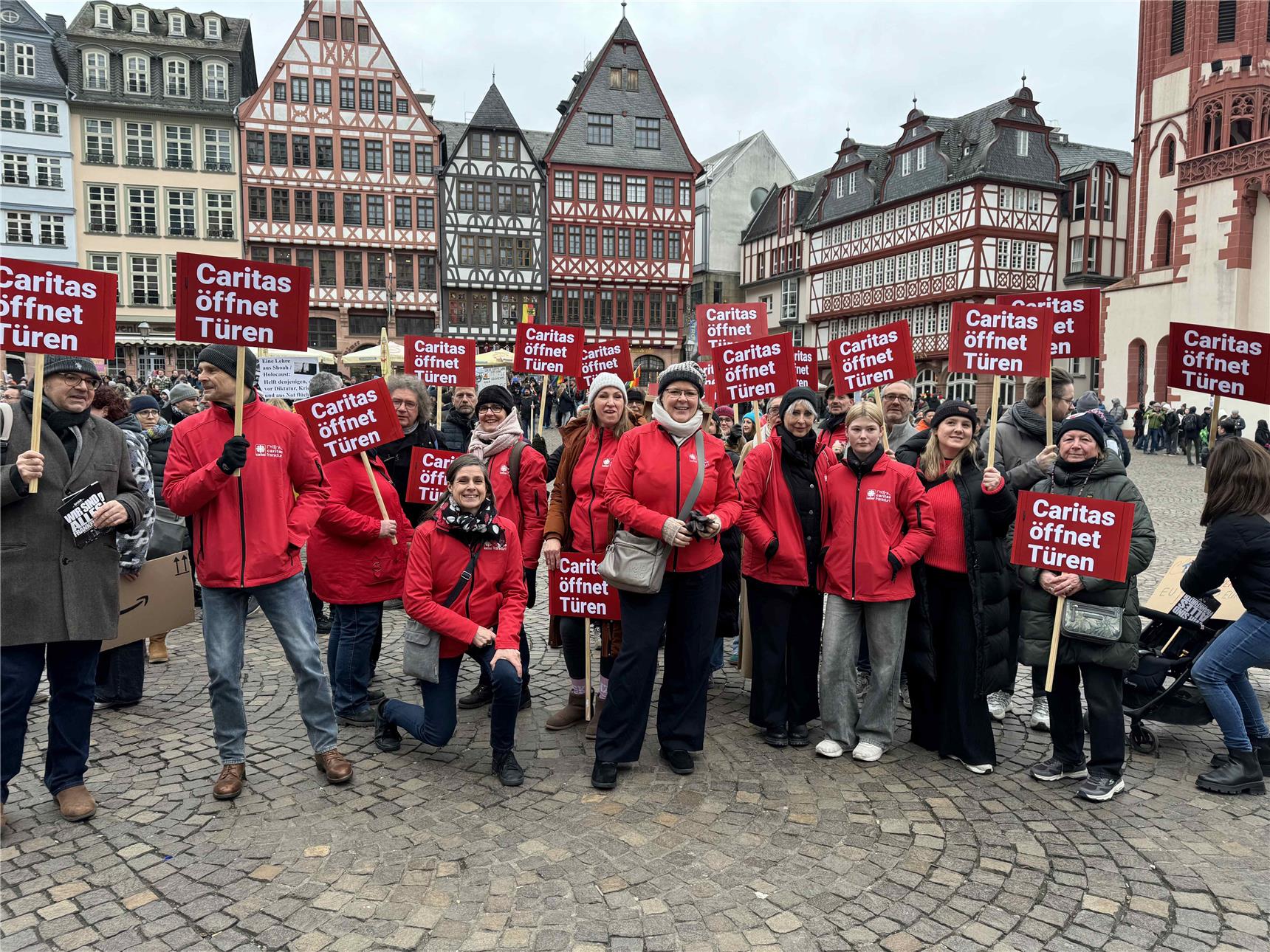 Die Teilnehmer'innen der Demo, ca. 20 Leute, tragen alle rot und halten rote Schilder in die Höhe mit der AUfschrift "Caritas öffnet Türen"