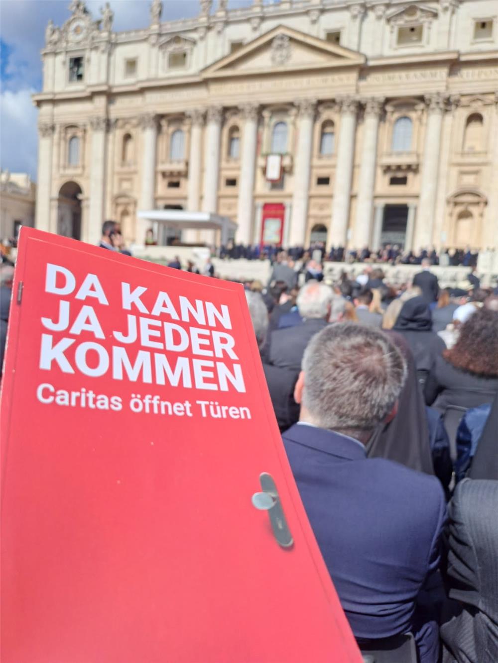 Im Vordergrund eine unscharfe rote Kampagnentür, im Hintergrund der mit Menschen gefüllte Petersplatz-