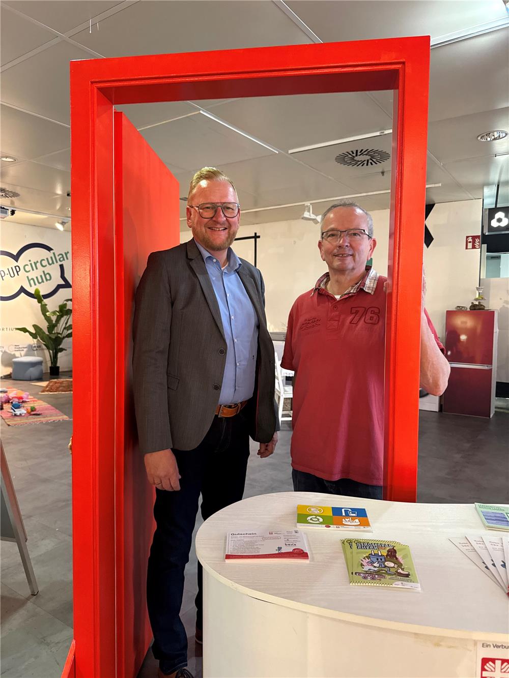 Matthias Timmermann (Caritas Direktor) und Christoph Dreger (Leitung des Stromspar Checks) stehen im Pop up Store des Stromsparchecks in einem roten Türrahmen, der Caritas Tür.