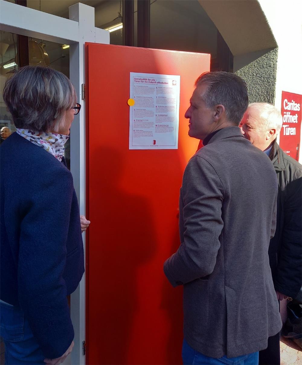 Thorsten Frei und E. M. Welskop Deffaa vor einer roten Tür, an der die 10 Thesen des Deutschen Caritasverbands zur Sozialpolitik hängen.