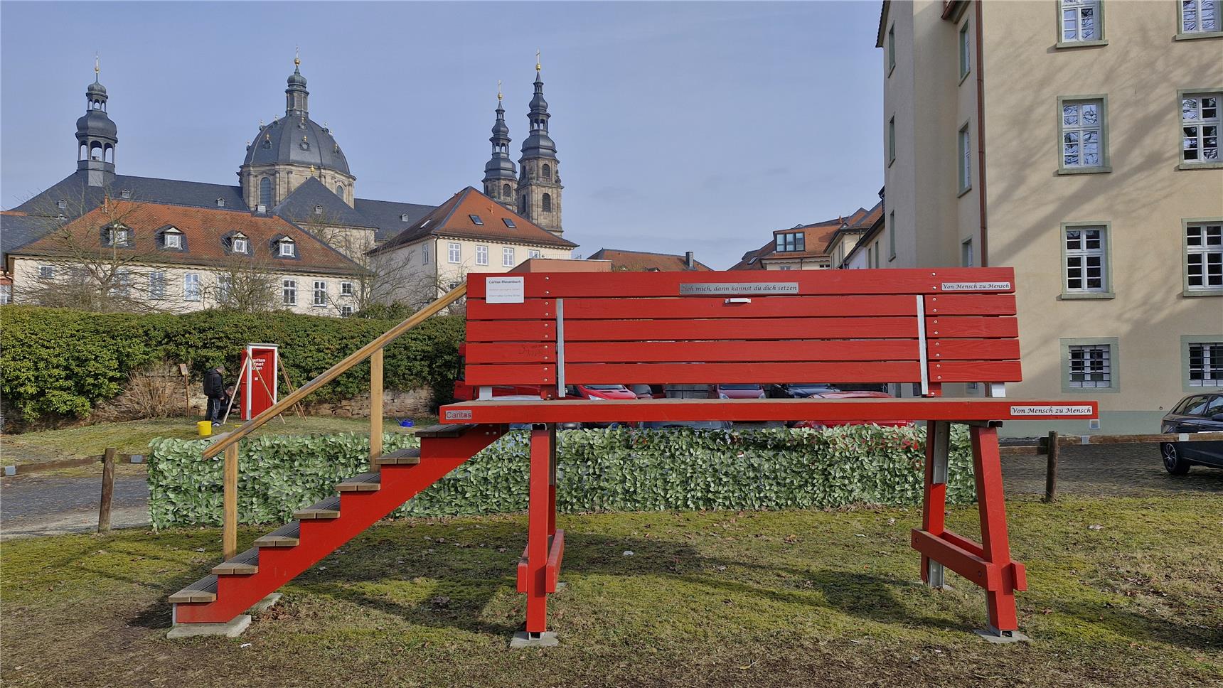 Eine große rote Riesen-Bank steht im Hof des Caritas-Hauses Wilhelmstraße. Im Hintergrund ist die Tür zu sehen.