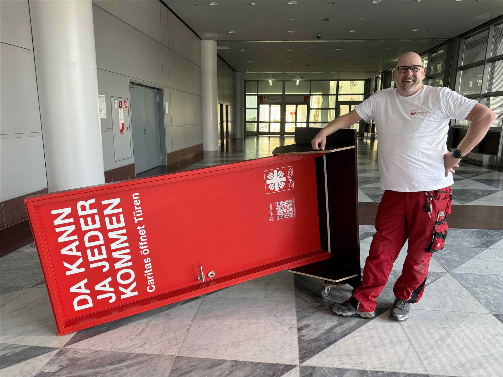 Hausmeister Manolito Haub der Caritas Frankfurt mit der transportabel roten Tür, welche gekippt auf einem Rollwagen liegt..