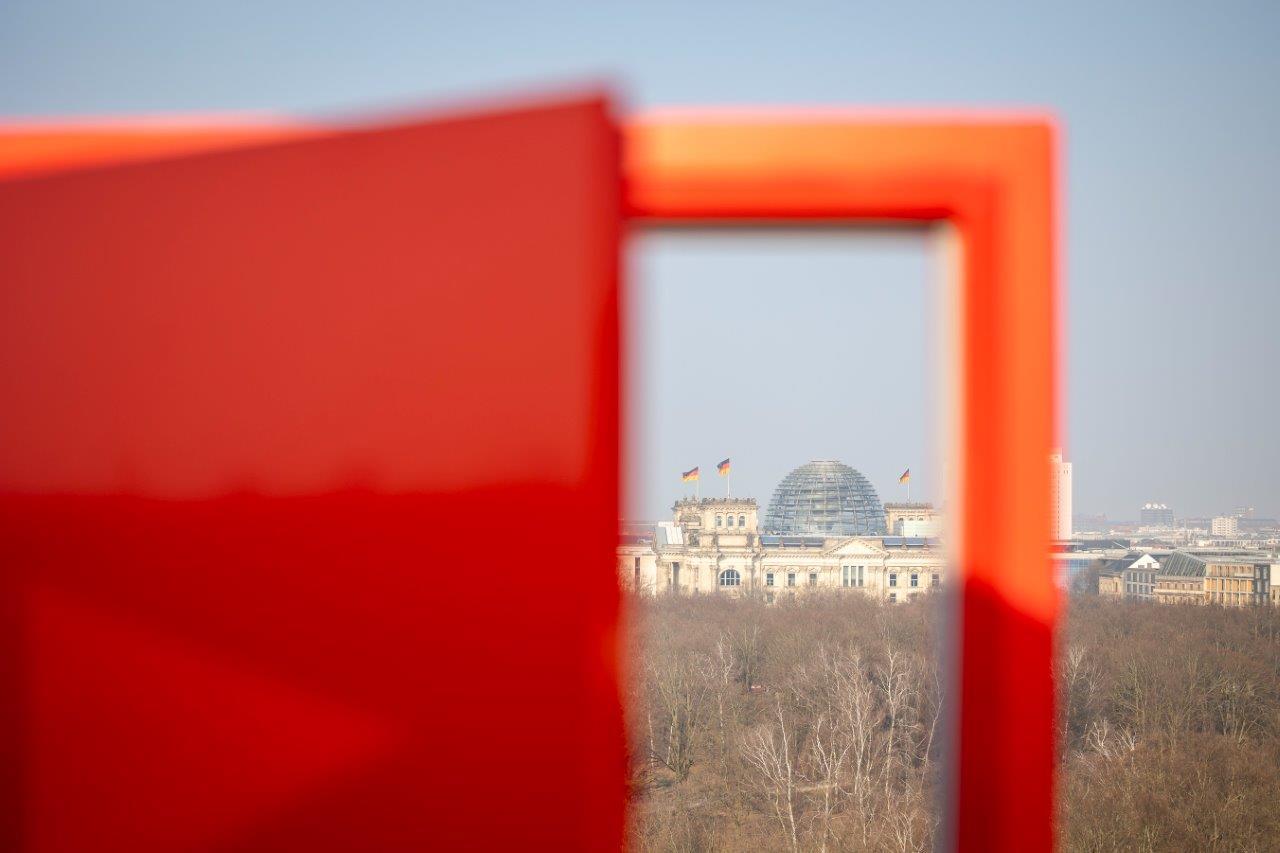 Blick durch den unscharfen Rahmen einer roten Kampagnentür in die Ferne auf das Bundestagsgebäude in Berlin. 
