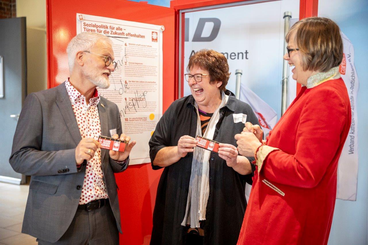 Der Geschäftsführer des VKAD, Andreas Wedeking (l), die Vorsitzende des VKAD, Barbara Dietrich-Schleicher und die Caritas-Präsidentin, Eva Maria Welskop-Deffaa unterhalten sich lachend vor einer roten Kampagnentür, an der die 10 Thesen 