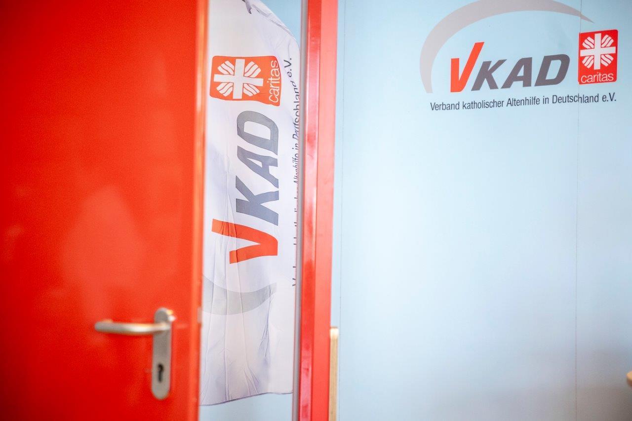 Durch die rote Kampagnentür ist eine weiße Flagge mit dem VKAD-Logo und dem Caritas-Flammenkreuz zu sehen. 