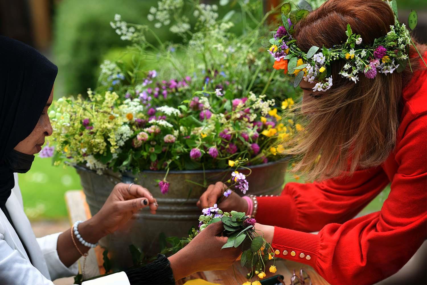 Eine Frau mit Kopftuch und eine Frau ohne Kopftuch knüpfen Blumenkränze.