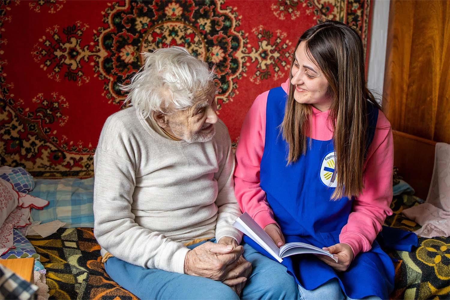 Ein älterer Herr und eine Frau, die eine Weste mit dem Logo der Caritas Ukraine trägt, sitzen auf einem Sofa.