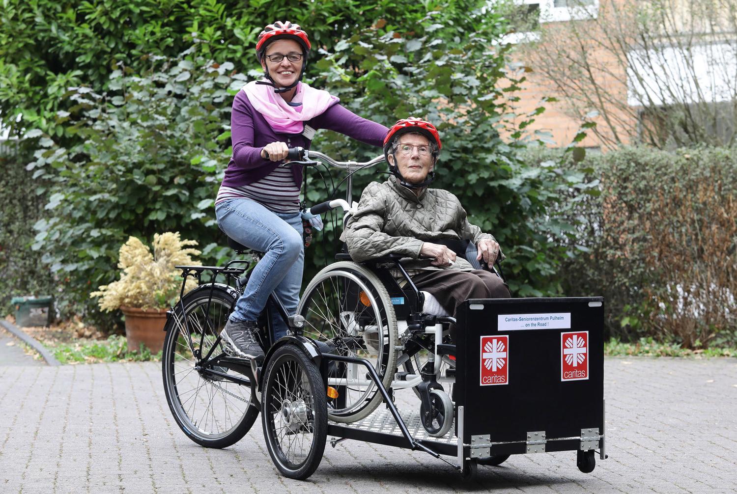 Ein Transport-Fahrrad ohne Motor wird von einer Caritas-Mitarbeiterin gefahren. Vorn im Cargobereich des Rades sitzt  eine ältere Bewohnerin in ihrem Rollstuhl..