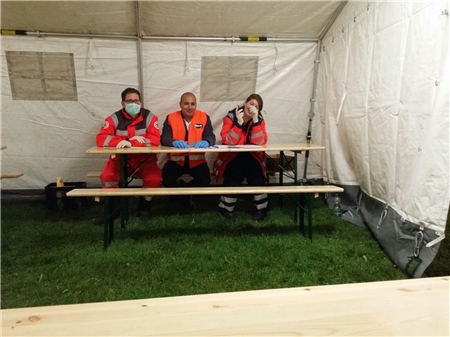 Zwei Mitarbeiter des Deutschen Roten Kreuzes sitzen gemeinsam mit einem Herrn an einer Bierzeltgarnitur in einem Rettungszelt.