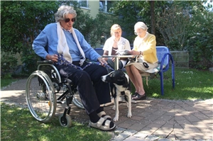Ältere Dame in Rollstuhl mit Hund. Im Hintergrund: Ältere Dame mit Mädchen auf Parkbank.