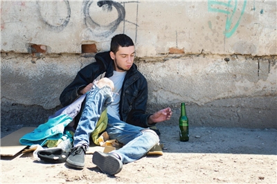Junger Mann, der auf der Straße auf Pappkarton sitzt, greift nach einer Bierflasche.