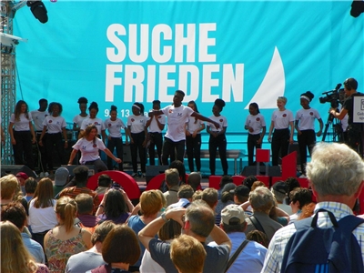Eine Bühne ist zu sehen, auf der junge Menschen unterschiedlicher Hautfarbe, alle in weißen T-Shirts, etwas vorführen. Im Hintergrund steht auf einem großen Transparent der Spruch "Suche Frieden"