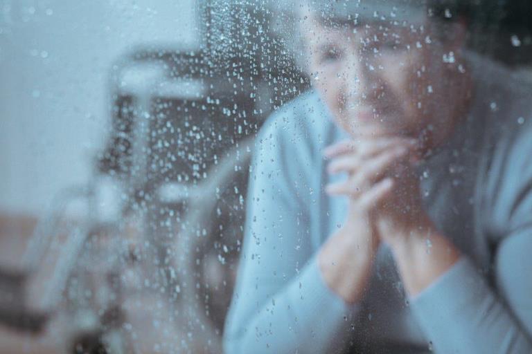 Eine alte Frau sitzt nachdenklich hinter einem Fenster mit Regentropfen.