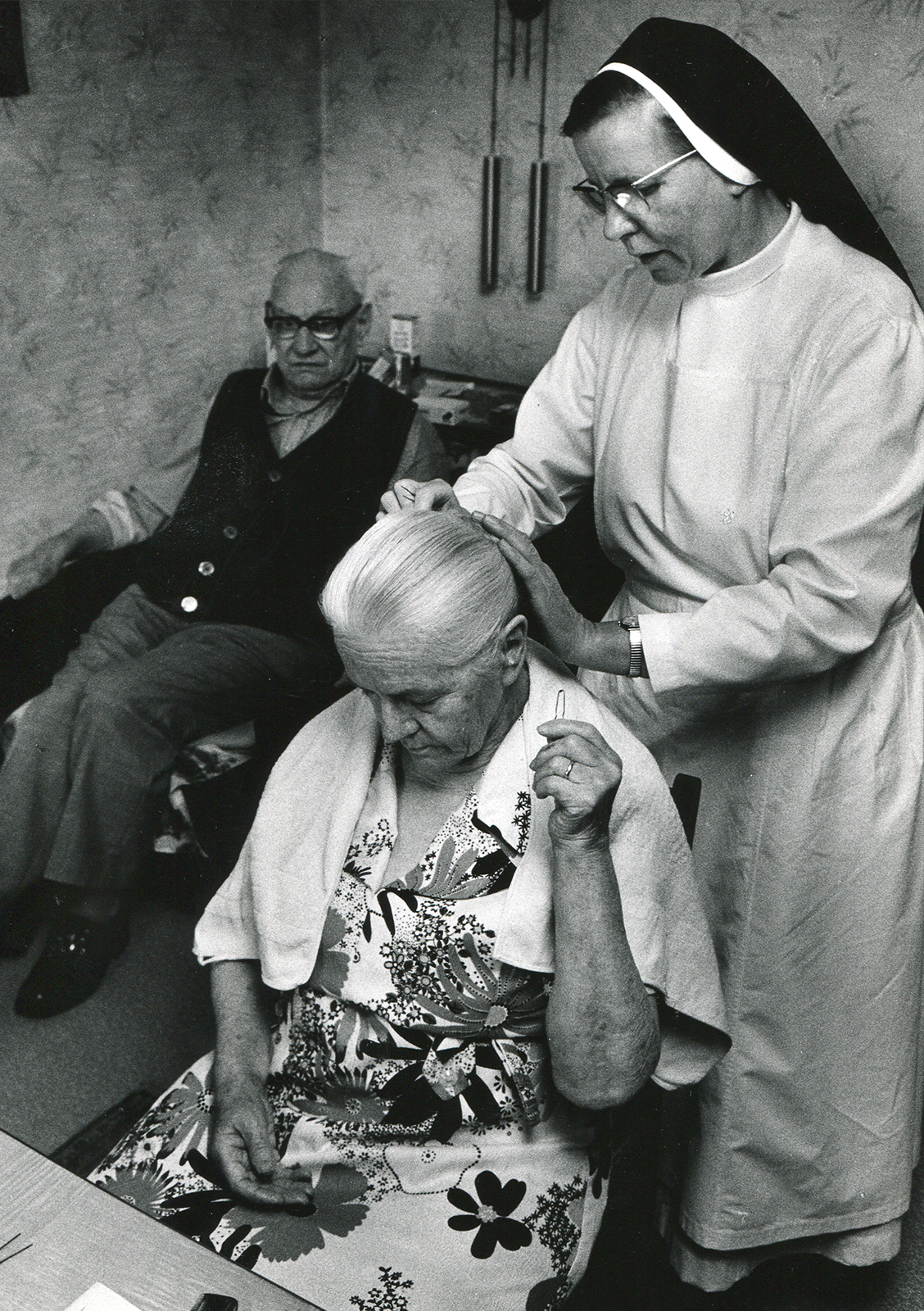 Eine ältere Dame und ein älterer Herr sitzen. Ordensschwester steckt ihr die Frisuer