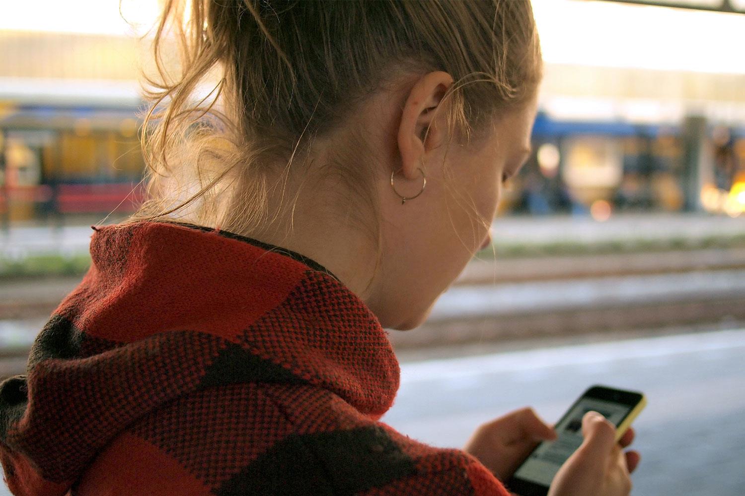 Eine junge Frau steht am Bahnsteig und Blickt auf ihr Smartphone