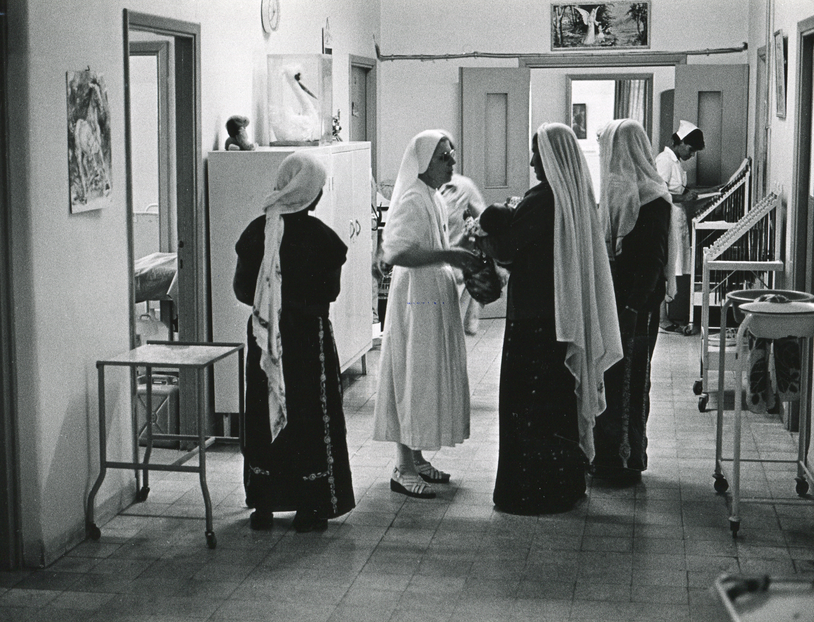 Im Flur stehen vier Frauen, Drei davon sind komplett im schwarz gekleidet und eine im weiß. Alle tragen einen langen Schleier. Sie haben Babies in der Hand.