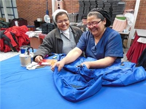 Frau bearbeitet ein Kleidungsstück, Ordensschwester schaut ihr über die Schulter