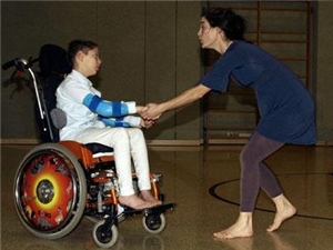 Frau nimmt im Rollstuhl sitzendes Kind an der Hand und führt es durch Turnhalle