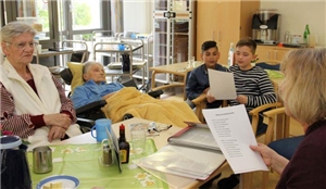 Senioren, Kinder und Betreuer sitzen am Tisch und singen gemeinsam. Einige von ihnen halten ein Liedblatt vor sich.