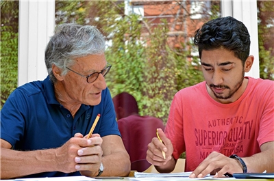 Ein älterer Mann sitzt mit einem jüngeren am Schreibtisch mit Stiften in den Händen.