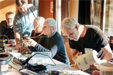 Vier ehrenamtliche Senioren an einem langen Werktisch prüfen Elektrogeräte.
