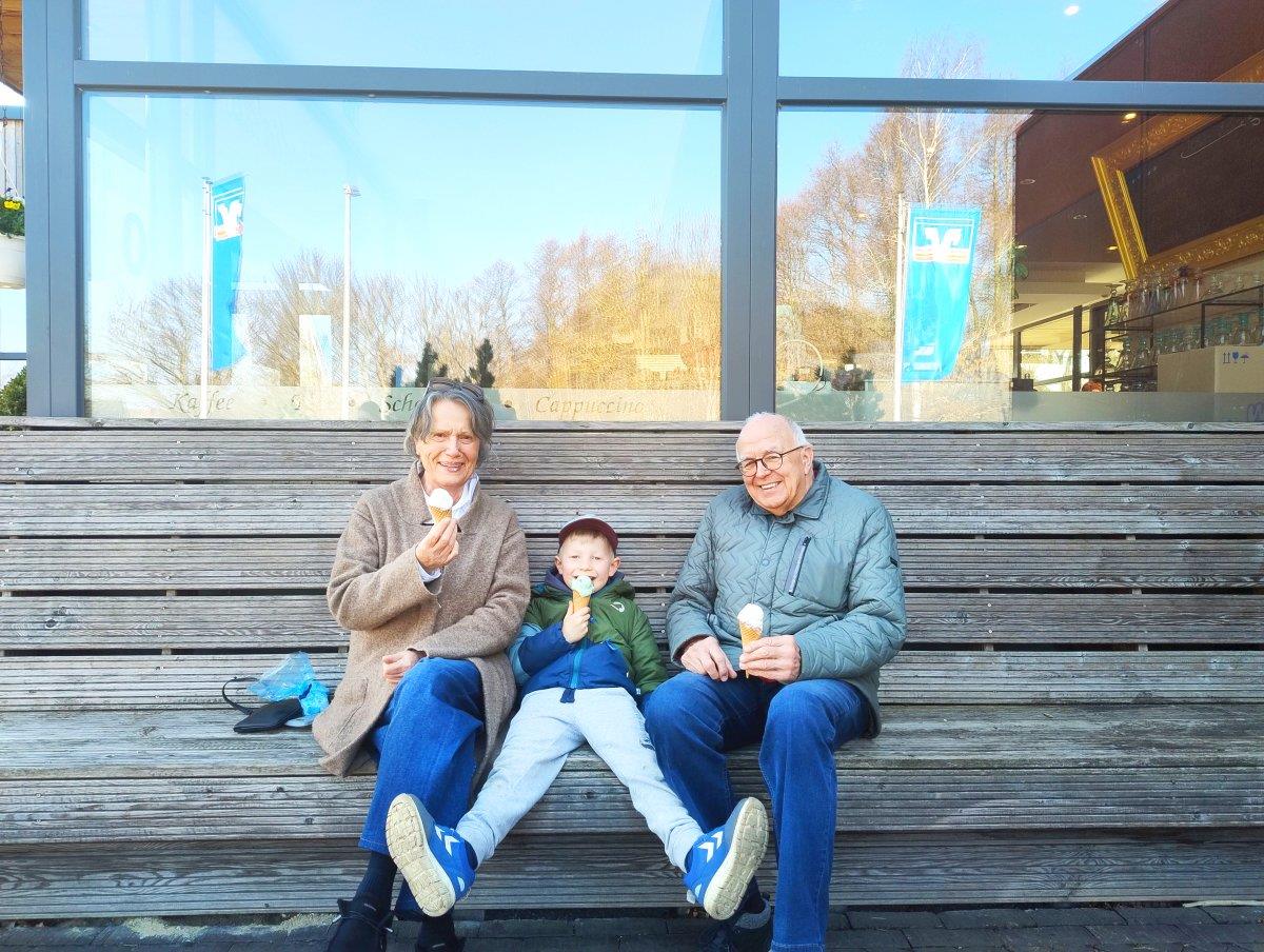 Ein älteres Paar sitzt mit einem Jungen auf einer Holzbank. Alle essen ein Eis. 