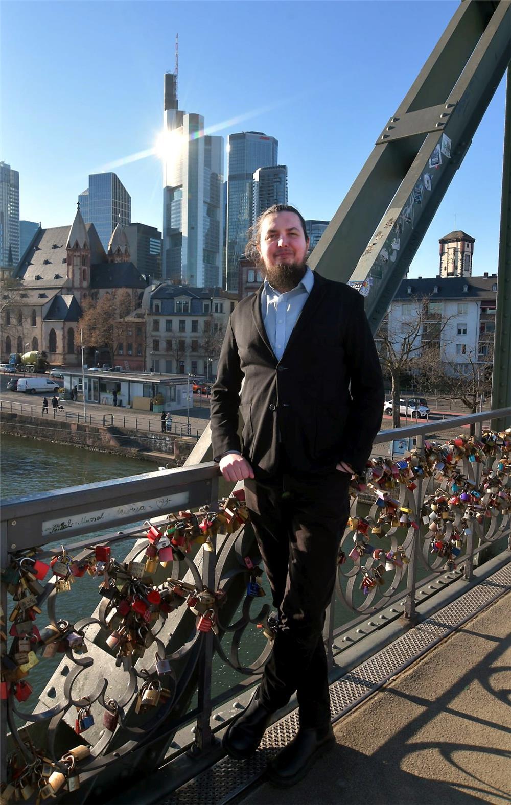 Ein Herr mit Zopf und Bart steht auf einer Brücke voller Liebesschlösser vor der Skyline von Frankfurt.