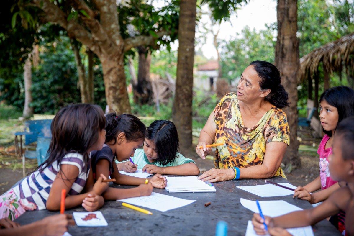 Kinder und Lehrerin sitzen im Amazonas mit Papier und Stiften um einen Tisch zwischen Palmen.