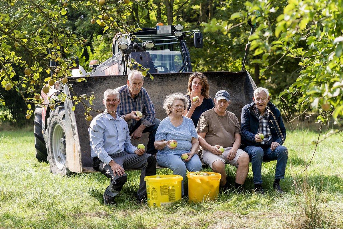 Zwei Frauen und vier Männer mit Äpfeln in der Hand sitzen auf der Schaufel eines Traktors.