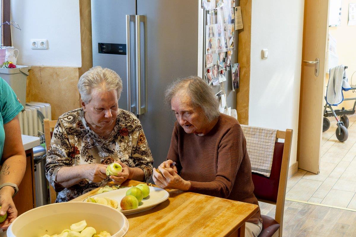 Zwei alte Bewohnerinnen des Pflegebauernhofs sitzen an einem Holztisch und schälen Äpfel.