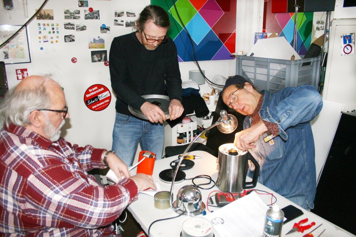 Drei Männer um einen im Tisch Repair-Café reparieren Wasserkocher und Lampen.