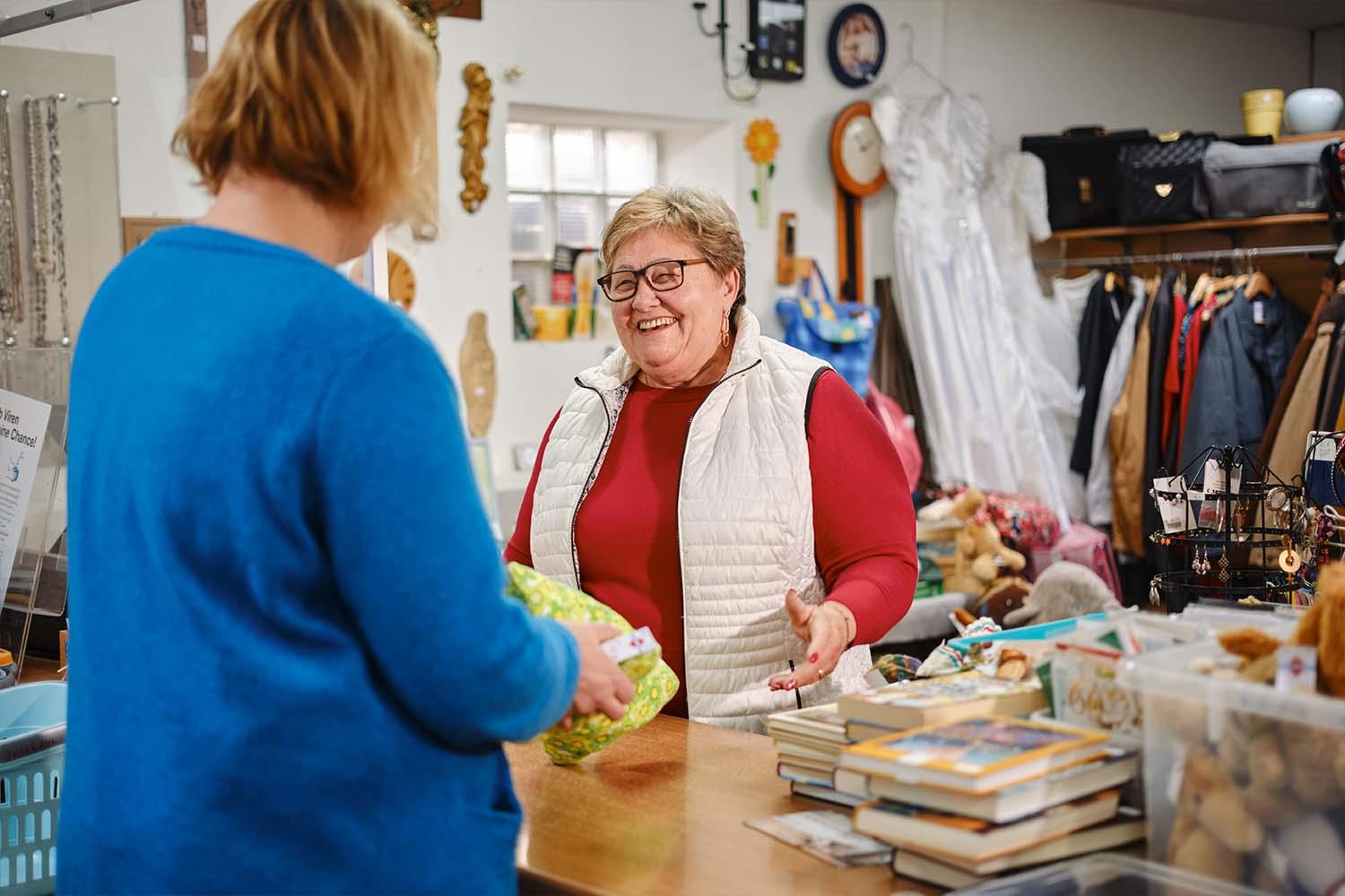 Ältere Dame mit kurzen blonden Haaren und Brille steht hinter einer Ladentheke und lächelt einer Frau zu.