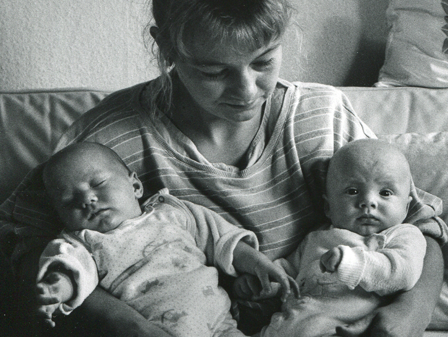 Eine junge Frau sitzt auf dem Sofa und hält zwei Babies in den Armen, ein baby schaut mit offenen Augen direkt in die Kamera, anderes Baby hat Augen zu.