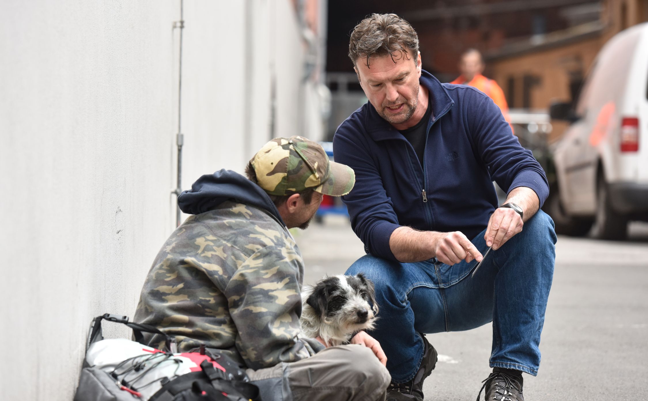 Ein Mann sitzt auf der Straße. Neben ihm steht ein Hund und ein Mann kniet neben ihm und spricht mit dem Mann.