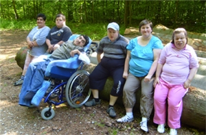 Eine Gruppe schwerstbehinderter Menschen sitzt auf einen gro�en dicken Baumstamm am Rande eines Wald