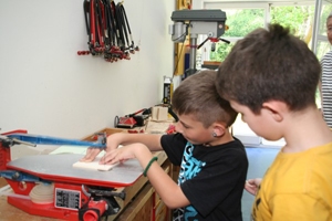 Zu sehen sind zwei Jungen, welche an einer S�ge arbeiten. Ein Junge s�gt seine Holzplatte gerade pr�zise aus. Der andere schaut ihm dabei zu.