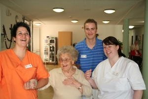 Eine Pflegerin im orangen Polo-shirt geht mit einer Seniorin spazieren. Zwei weitere Pflegepersonen folgen ihnen.