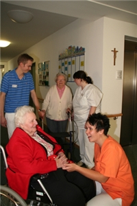 Die Pflegerin im orangen Polo-Shirt lacht mit eine Frau im Rollstuhl. Im Hintergrund sind zwei Pflegekräfte mit eine Bewohnerin