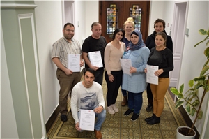 Seminarleitung Anne Geerken (hinten Mitte) mit Kollegin Farha Betto (davor) den Teilnehmenden und einer Referentin Kerstin Bender (hinten re.; Verbraucherzentrale NRW). Eine Gruppe von 8 Menschen. 5 Migranten halten stolz ihre Zertifikate in die Kamera.