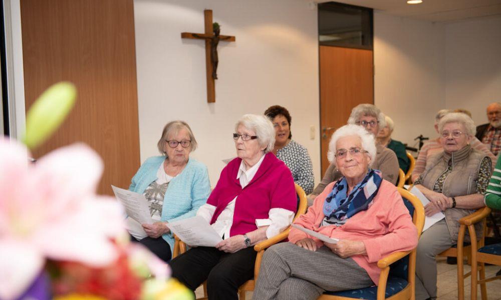 Seniorinnen sitzen im Gemeinschaftsraum mit Kreuz an der Wand