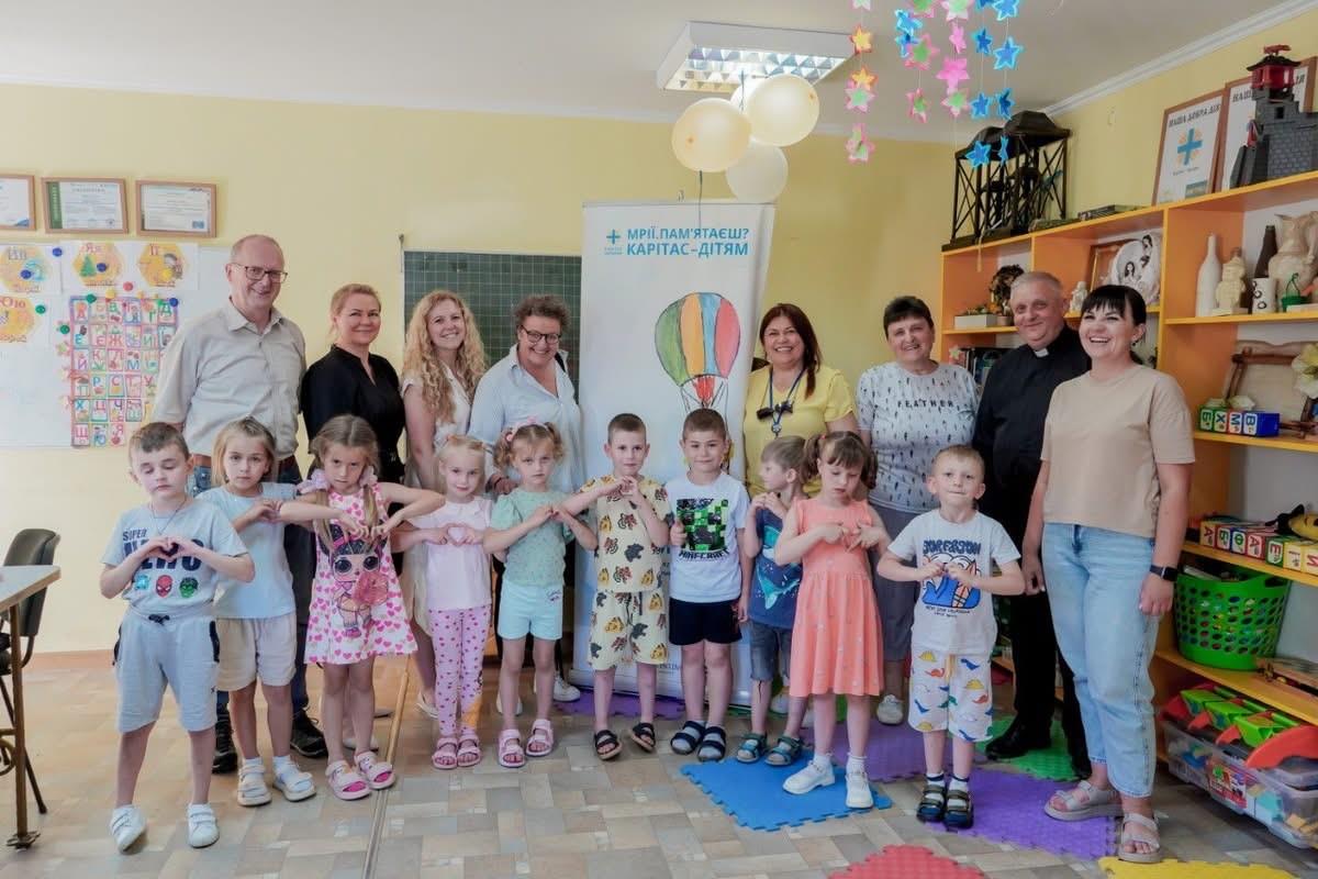 Ein Gruppenfoto: Im Vordergrund stehen zehn Kinder nebeneinander, dahinter haben sich acht Erwachsene aufgereiht. Neben der Gruppe ist ein Schrank mit vielen Spielzeugen zu sehen. Im Hintergrund befindet sich eine große Schreibtafel.