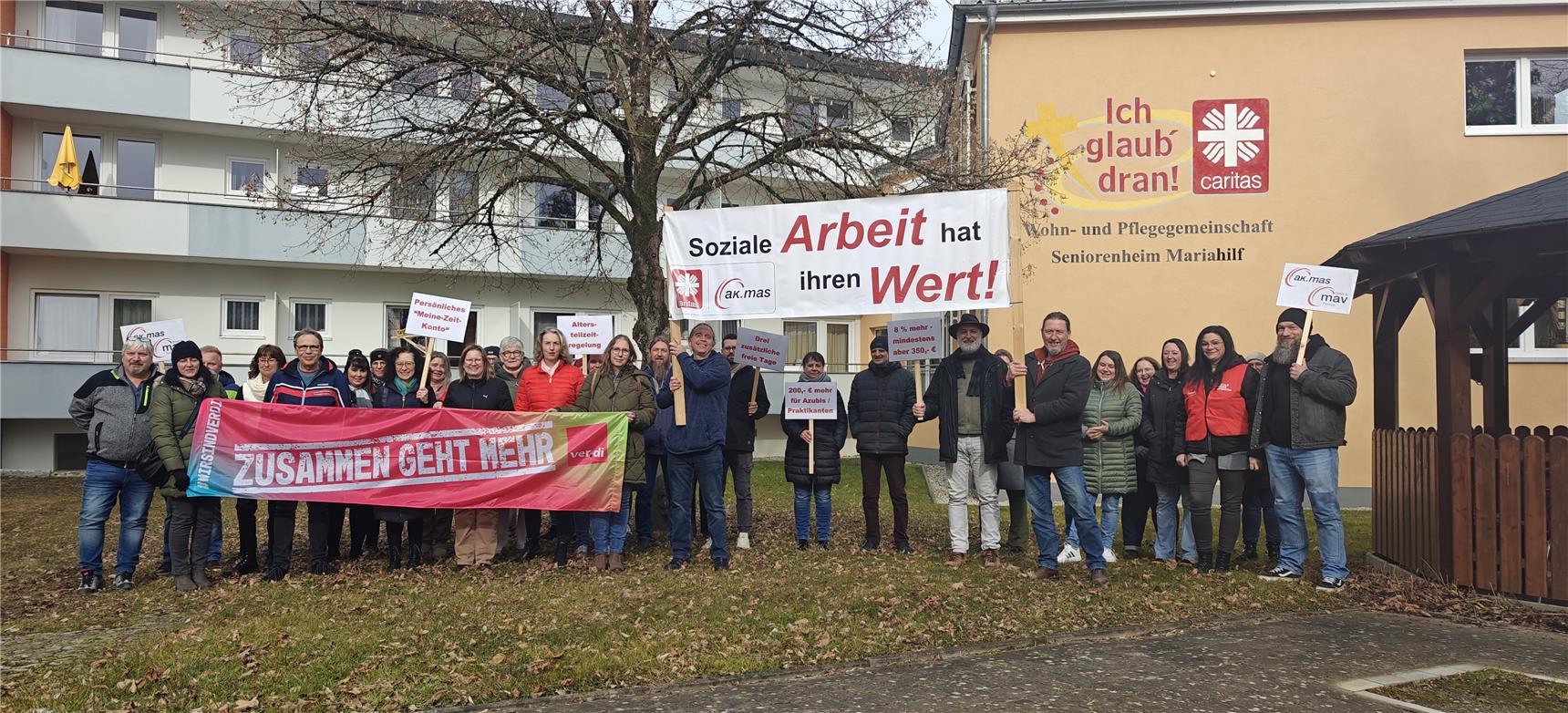 Eine Gruppe von Menschen protestiert vor dem Seniorenheim Mariahilf mit Bannern und Schildern für bessere Arbeitsbedingungen in der sozialen Arbeit, darunter Forderungen nach mehr Lohn.