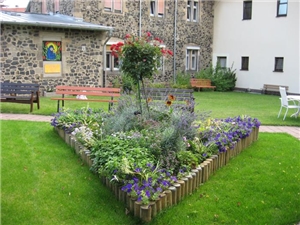 In einem dreieckig gestalteten Blumenbeet vor dem historischen Teil des Altenpflegeheimes blühen blaue, weiße und rote Blumen und Sträucher. Der alte Teil  des Gebäudes ist ein Mauerwerk aus blauen Ba