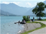Bad_Wiessee Wir blicken auf einen sauberen Bergsee. Rechts ist eine Bucht auf der Menschen sich zum baden vorbereiten. Auf einer schattigen Bank unter einem großen Baum sitzt jemand, der auf den See blickt. Ein K
