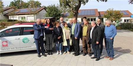Das Pressefoto entstand während des Besuches der Grünen in Fulda-Maberzell: Die Gruppe von Grünen- und Caritas-Vertretern mit Elektro-Fahrzeug und vor den Caritas-Häusern mit Stromerzeugung.  