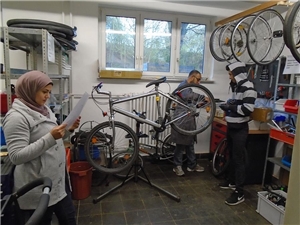 Ein Fahrrad wird auf einem Ständer repariert. Regale und Werkbänke mit Werkzeug und Material sind an den Wänden zu sehen.